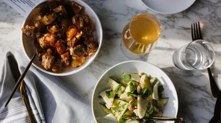 Two bowls sit on a table. One is filled with seasoned beef chunks and the other is filled with a sad with wedges of parmesan cheese. There is a glass of white wine between them. 