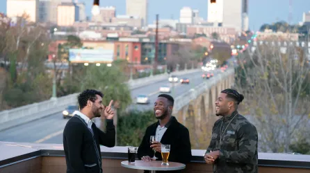 Three men enjoy drinks on a rooftop bar overlooking the city of Columbia SC at sunset.