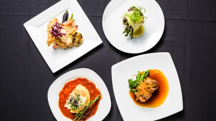 four plates are laid next to one other on a black table cloth. specially prepared food sits on top of each plate.