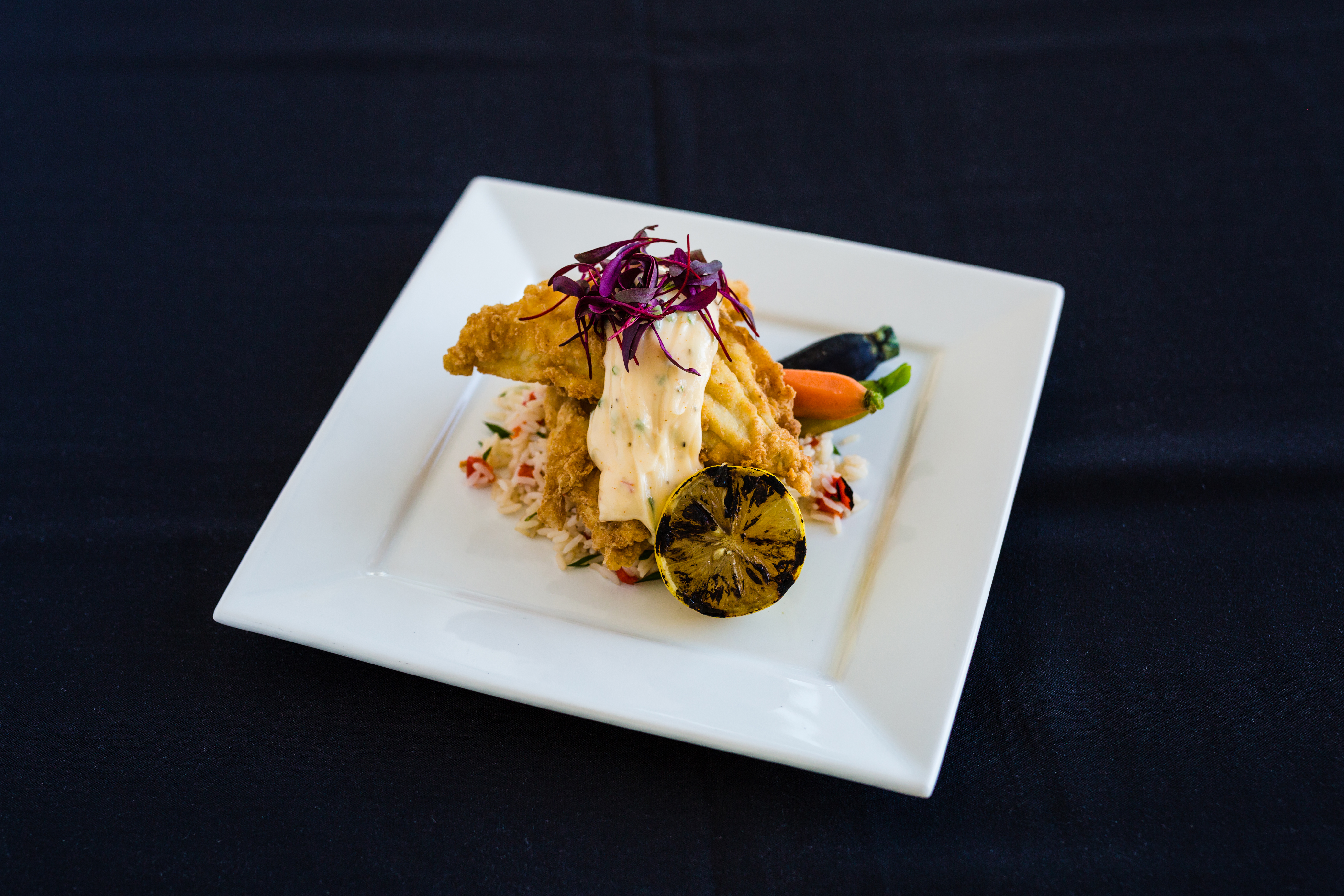 A white square plate sits on a black table cloth. Fried fish is on the plate on top of a bed of gratin potatoes. There is a grilled, blackened lemon on the forefront of the image.